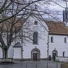 Eine romanische Kirche in Seitenansicht mit heller Fassade, rot eingedecktem Dach und Glockenturm.