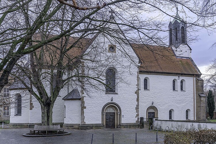 Eine romanische Kirche in Seitenansicht mit heller Fassade, rot eingedecktem Dach und Glockenturm.