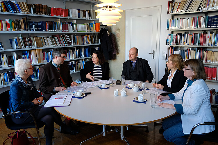 Sechs Personen sitzen um einen runden Tisch herum, im Hintergrund sind Bücherregale zu sehen.