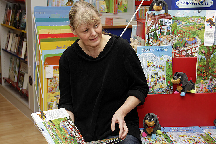 Eine Frau sitzt und schaut in ein aufgeschlagenes Buch mit farbigen Illustrationen.