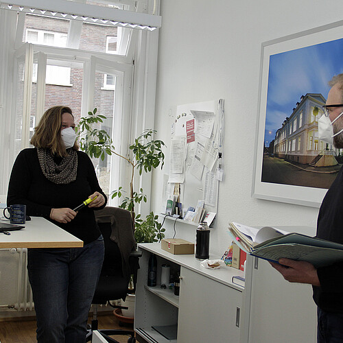 Eine Frau und ein Mann stehen in einem Büro und unterhalten sich, beide tragen FFP3-Schutzmasken.