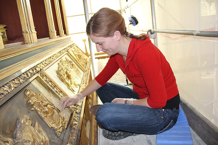 Eine junge Frau sitzt im Schneidersitz auf einem Gerüst vor einem vergoldeten Altar und bearbeitet die Oberfläche.