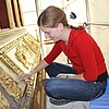 Eine junge Frau sitzt im Schneidersitz auf einem Gerüst vor einem vergoldeten Altar und bearbeitet die Oberfläche.