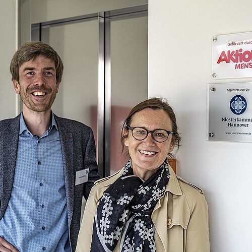 Zwei Personen, Johannes König und Silke Sackmann, stehen vor einer Eingangstür. Neben ihnen an der wand sind zwei Schilder angebracht, die auf die Förderung der Aktion Mensch und der Klosterkammer Hannover hinweisen.