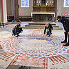 Drei Frauen und ein Mann stehen oder knien auf einem einem mit großen Mustern verzierten Boden und schauen auf die restaurierten Elemente.