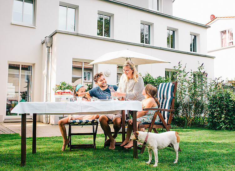 Eine Familie mit zwei Töchtern sitzt an einem Tisch im Garten, daneben sitzt ein Hund. Im Hintergund ist eine weiße Häuserfassade zu sehen.