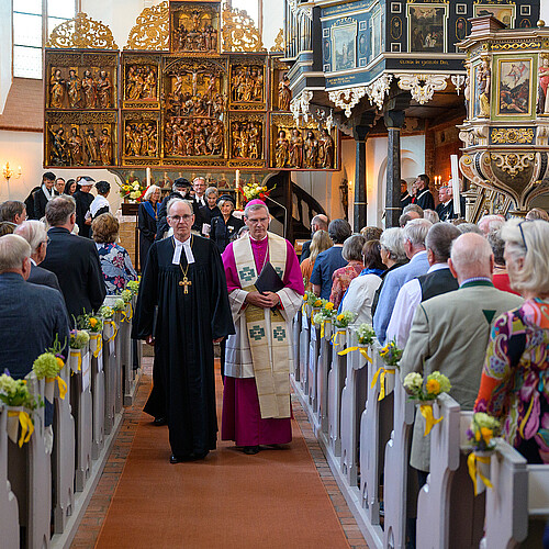 In der festlich geschmückten Klosterkirche Lüne schreiten zwei Männer in liturgischer Kleidung durch den Mittelgang. Die Bankreihen sind durchgängig besetzt.