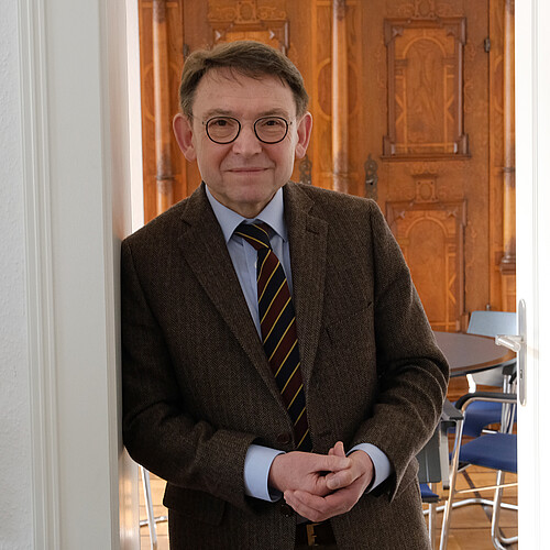 Ein Mann in braunem Sakko, mit Krawatte und Brille steht in einem Türrahmen.