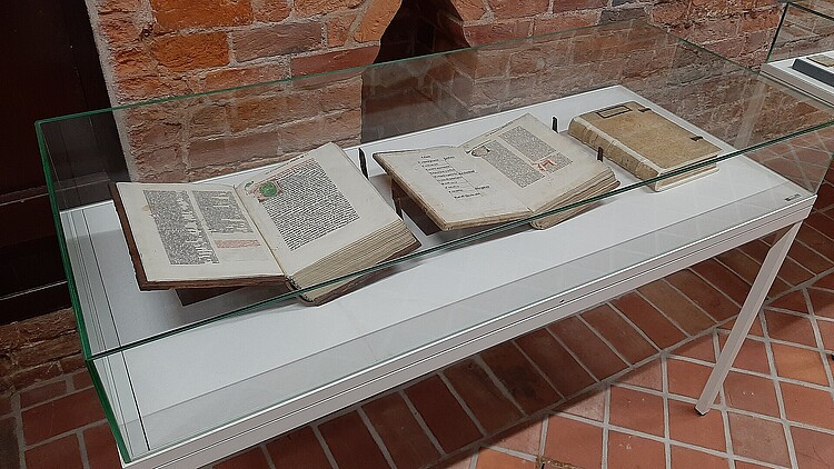 In einer Vitrine, die mit Glas abschließt, sind drei historische Bücher ausgestellt.