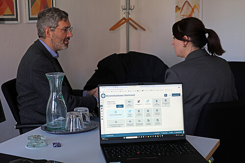 Im Vordergrund des Bildes ist ein Laptop, im Hintergrund sitzen zwei Personen.