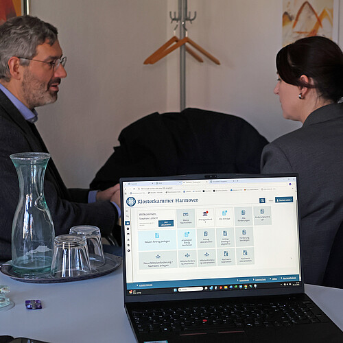 Im Vordergrund des Bildes ist ein Laptop, im Hintergrund sitzen zwei Personen.