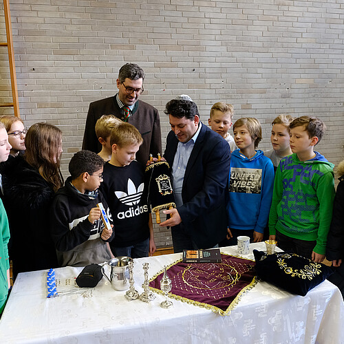 Eine Gruppe Schülerinnen und Schüler und zwei Erwachsene stehen um einen Tisch mit Gegenständen aus dem jüdischen Leben herum.