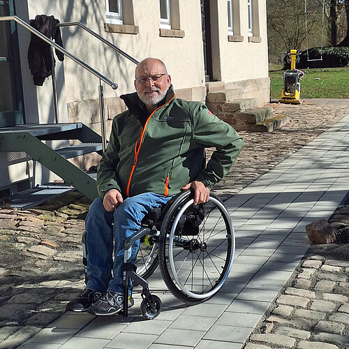 Ein Mann im Rollstuhl steht auf einem Weg neben einem Gebäude.