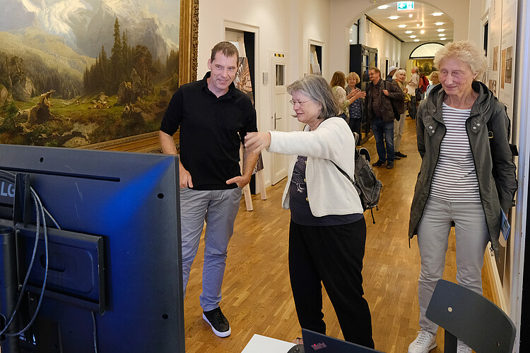 Besucher zeigen auf einen Monitor. Mitarbeiter erklärt etwas.