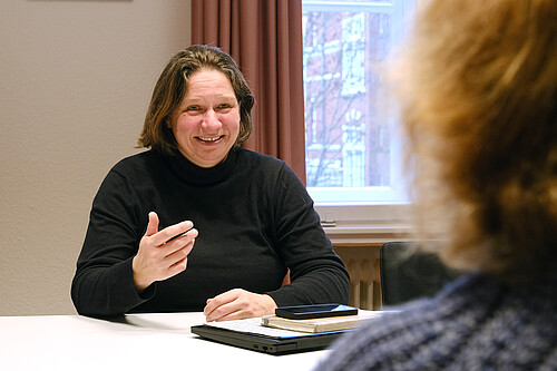 Eine Frau sitzt an einem Tisch und spricht mit einer anderen Person, die von hinten zu sehen ist.