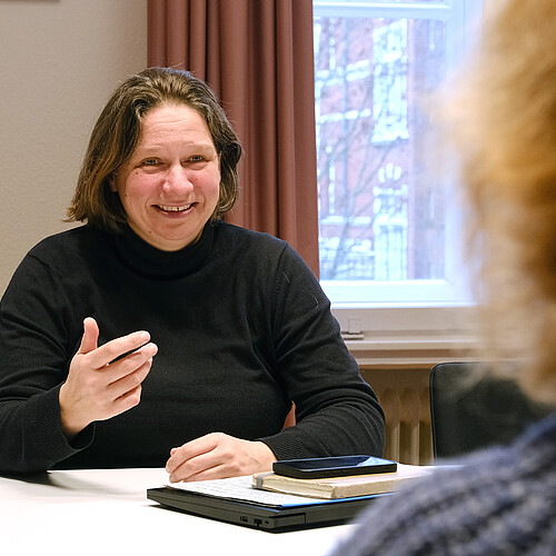 Eine Frau sitzt an einem Tisch und spricht mit einer anderen Person, die von hinten zu sehen ist.