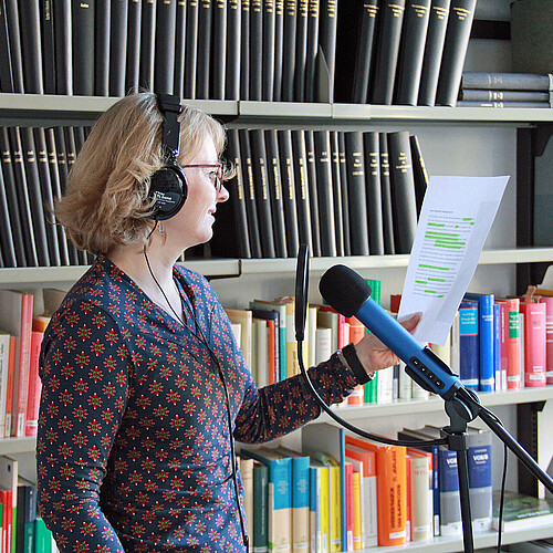 Eine Frau mit Kopfhörern steht vor einem Mikrofon und nimmt einen Text auf, im Hintergrund eine Bücherwand.