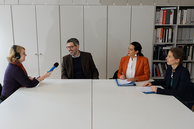 Frau mit Mikrofon und Kopfhörern interviewt einen Mann und zwei Frauen an einem großen Tisch