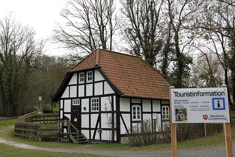 Ein kleines Fachwerkhaus - im Vordergrund ist ein Schild zu sehen, auf dem Informationen zur Touristinformation zu lesen sind - steht inmitten einer Parkanlage.