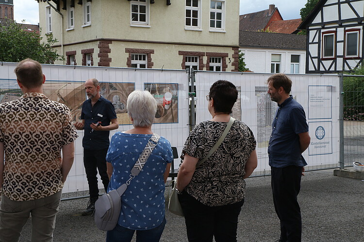 Mehrere Personen schauen sich Planen mit Illustrationen an einem Bauzaun an. Ein Mann erklärt etwas.