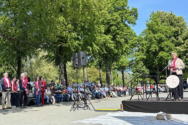 Eine Frau steht auf einem Podium und spricht, rundherum hören Menschen zu.