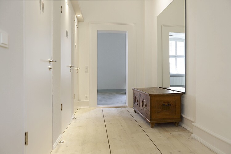 Zwei Türen zu WCs, davor heller Holzboden, eine alte Truhe und ein Spiegel.