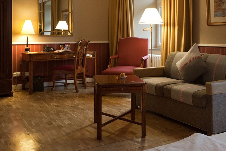 Ein edles, dunkel eingerichtetes Hotelzimmer mit einem Sofa, einem Sessel und einem antiken Schreibtisch.