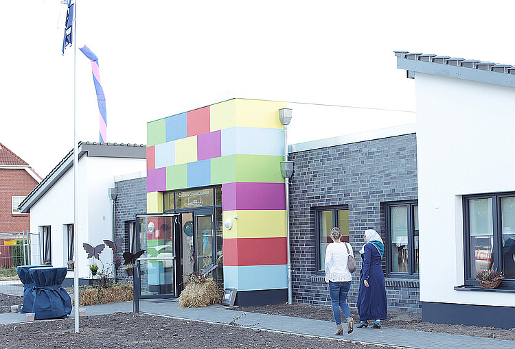 Vor dem Neubau des Kinderhospiz mit farbigen Elementen laufen zwei Frauen einen Weg entlant.