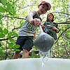 Ein Junge schöpft Wasser mit einer Kelle in einen Eimer, im Hintergrund sind Bäume zu sehen.