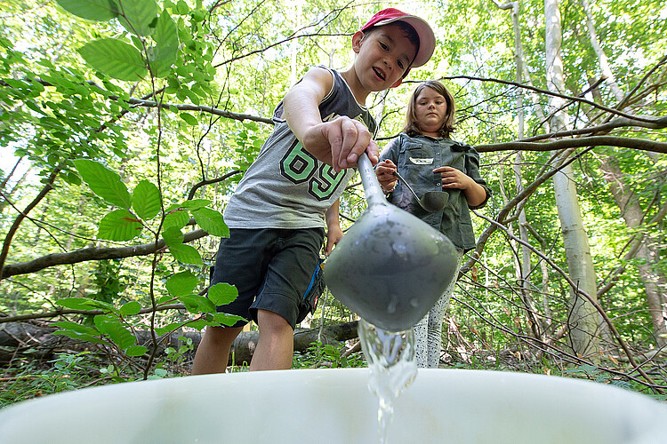 Ein Junge schöpft Wasser mit einer Kelle in einen Eimer, im Hintergrund sind Bäume zu sehen.