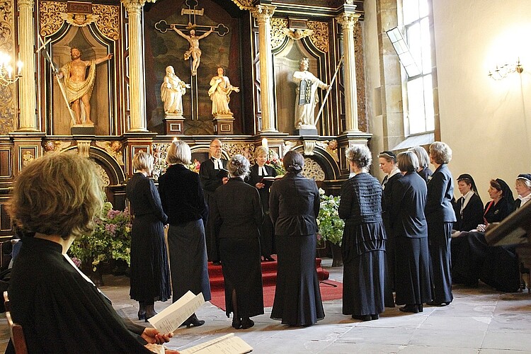 Neun Frauen in schwarzen Gewändern stehen im Halbrund vor einem Pastor in einer Kirche während eines Gottesdienstes.