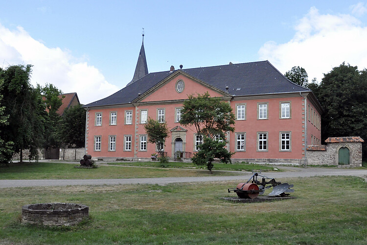 Leicht schräge Gebäudeansicht mit angeschnittener Mauer rechts und ein Stück des Kirchturms im Hintergrund links.