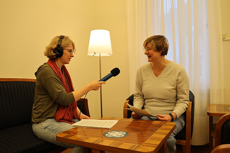 Zwei Frauen an einem Couchtisch, eine Frau mit Mikrofon und Kopfhörern interviewt die andere.