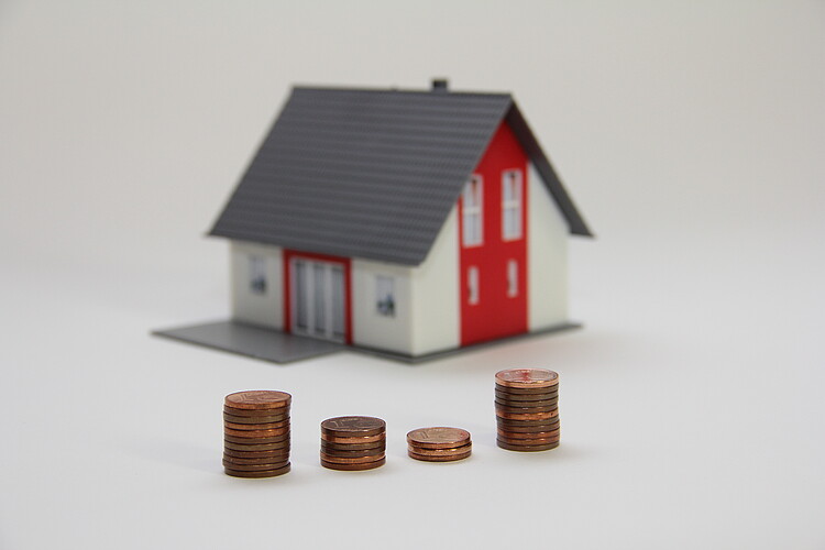 Symbolfoto für den Wertverfall: Im Vordergrund sind unterschiedlich hohe Stapel mit Cent-Stücken zu sehen, im Hintergrund steht ein kleines Modellhaus.