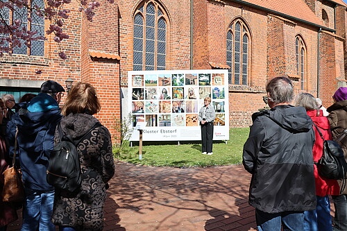 Eine Frau steht  auf einer Wiese vor einer Tafel mit 28 Fotos im Quadrat.