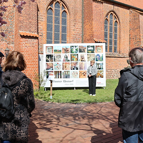 Eine Frau steht  auf einer Wiese vor einer Tafel mit 28 Fotos im Quadrat.
