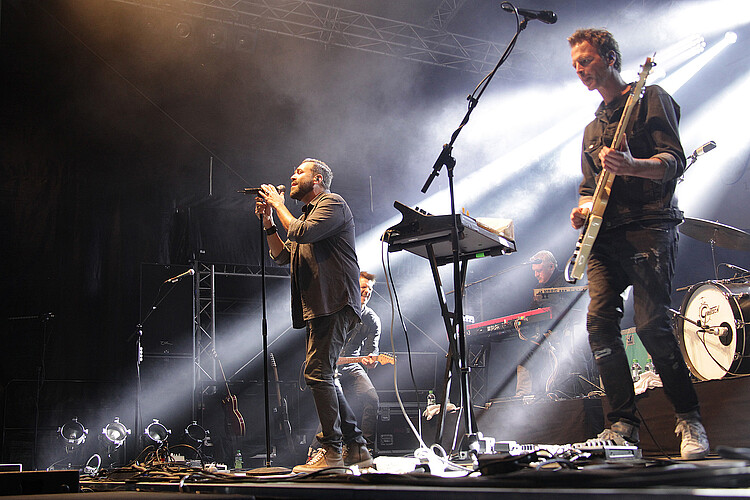 Auf einer Bühne steht ein Sänger an einem Mikrofon, daneben sind zwei weitere Musiker zu erkennen.
