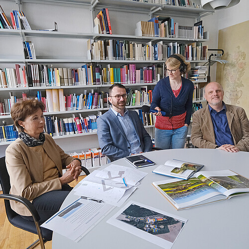 Drei Personen sitzen an einem Tisch, eine Person steht mit einem Mikrofon dazwischen.