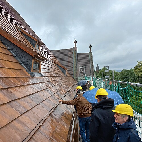 Mehrere Personen mit Bauhelmen und Regenschirmen bei Regen auf einem Baugerüst vor einem frisch gedeckten Kirchendach.
