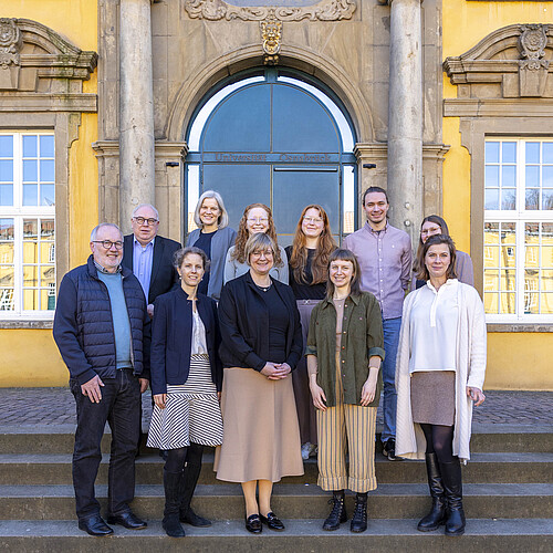 Elf Personen stehen auf einer Treppe vor einem Gebäude mit gelber Fassade.