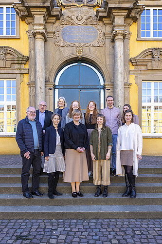 Elf Personen stehen auf einer Treppe vor einem Gebäude mit gelber Fassade.