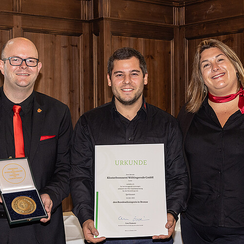 Drei Männer und zwei Frauen nebeneinander. Ein Mann hält eine Schachtel mit einer Medaille. Ein Mann hält eine Urkunde.