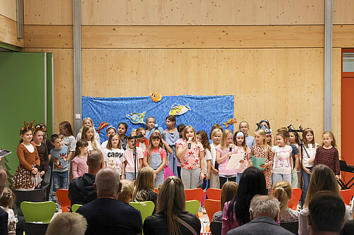 Eine Kindergruppe steht für eine Aufführung in einer Schulaula vor Publikum.