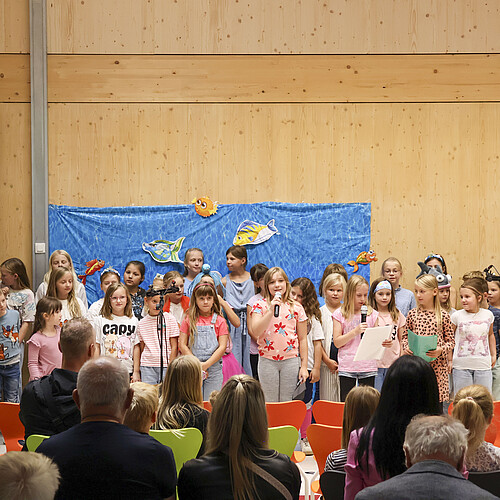 Eine Kindergruppe steht für eine Aufführung in einer Schulaula vor Publikum.