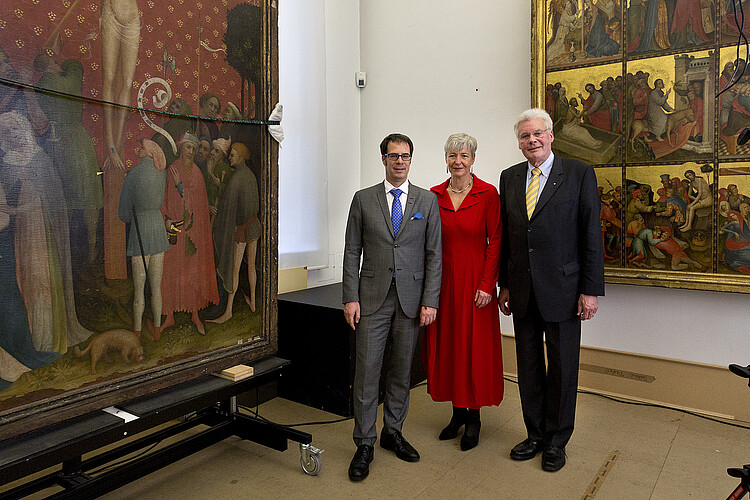 Drei Personen stehen vor historischen Bildern an dem Teil eines Altars, der teils vergoldet ist.
