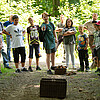 Eine Gruppe von Schulkindern steht im Wald in einem Halbkreis, ein Junge wirft einen Zapfen in einen Korb.