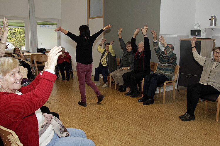 Seniorinnen und Senioren sitzen in einer Runde auf Stühlen und machen Bewegungsübungen mit den Armen.