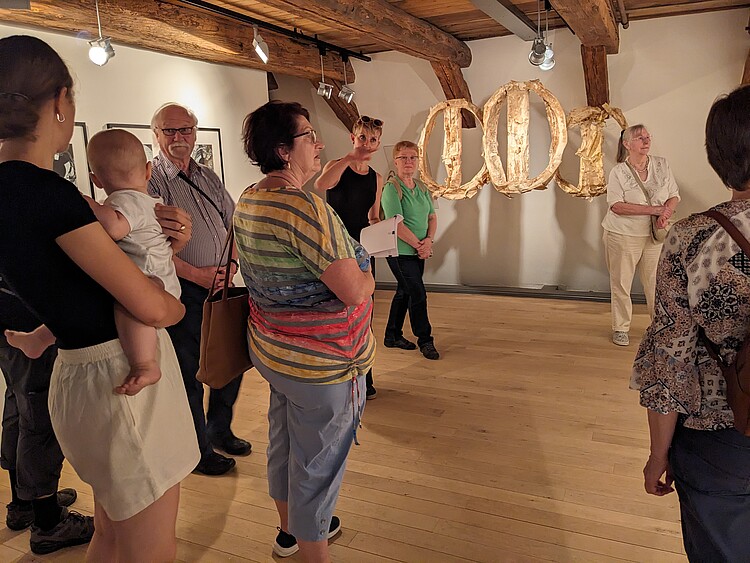 Mehrere Personen schauen sich im Dachgeschoss eines historischen Gebäudes eine Kunstausstellung an.