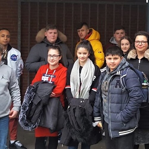 Eine Gruppe von Schülerinnen und Schülern steht zusammen mit einer Bildungsbegleiterin vom 1. Sinti-Verein Ostfriesland e.V. auf einem Schulhof.