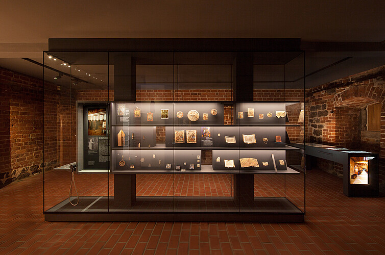 Eine große gläserne Vitrine steht in einem Museumsraum. Einzelne Objekte darin sind beleuchtet.
